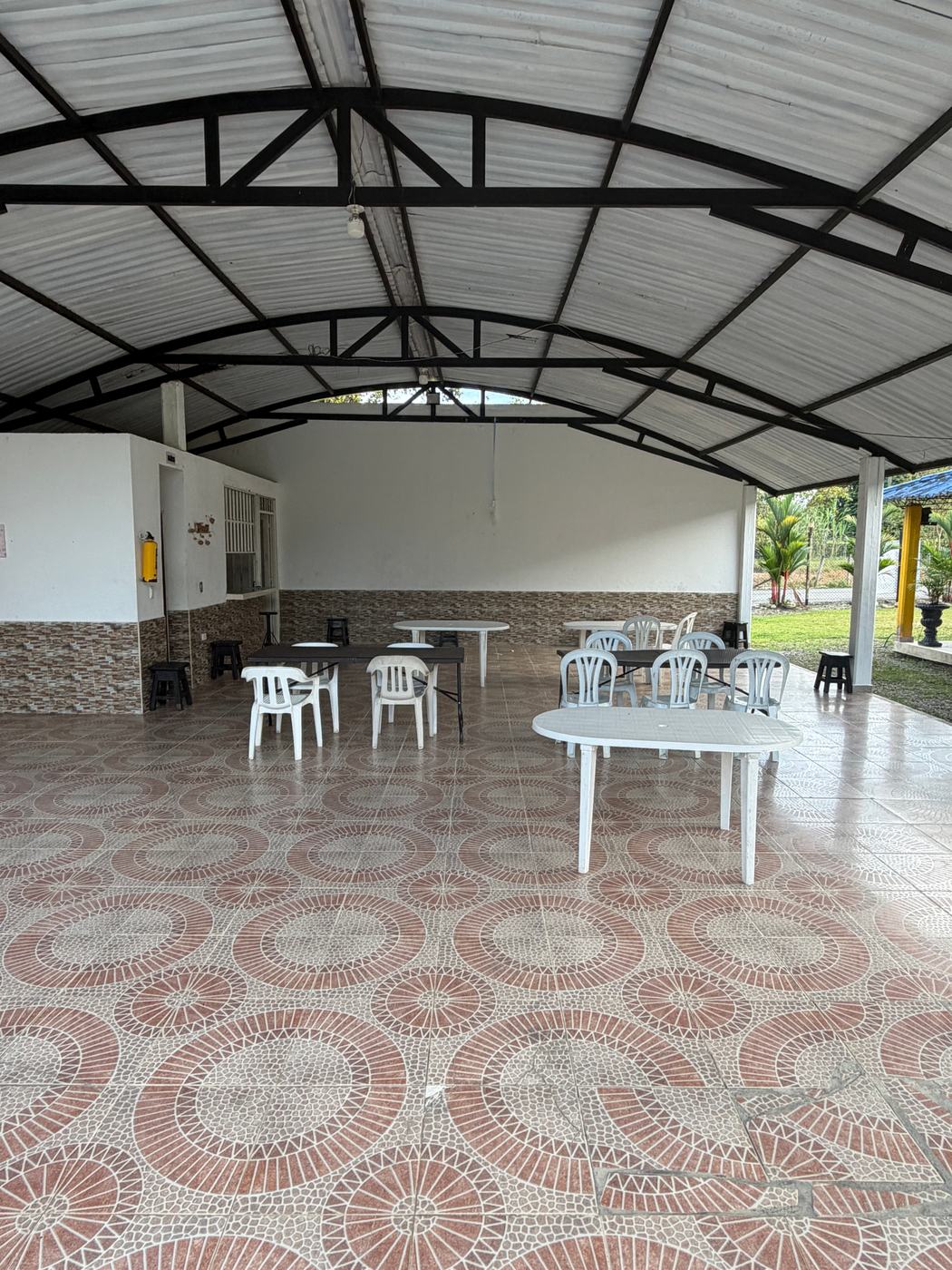 Salón social cubierto de techo alto, con piso de baldosa y mesas con sillas blancas